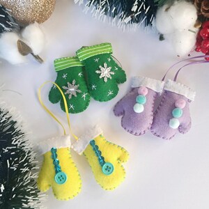 May include: Three pairs of felt mitten ornaments. One pair is green with a snowflake design, one pair is purple with white pom-poms, and one pair is yellow with a blue button.