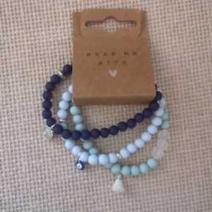 Peut inclure: Trois bracelets superposés avec des perles bleues, bleu clair et blanches. Les bracelets ont des breloques, dont un cercle argenté, un mauvais œil bleu et un pompon blanc.