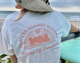 Puede incluir: Una camiseta blanca con el texto "SURFERS AGAINST SLAVERY" en semicírculo sobre las palabras "Waves Of Freedom". El diseño incluye una ilustración floral en color melocotón. La persona lleva un sombrero beige y sostiene una tabla de surf.