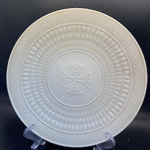 Könnte beinhalten: Weiße Keramik-Weihnachtsteller mit geprägten Mustern und einem zentralen Kreuzdesign. Der Teller trägt die Aufschrift "CHRISTMAS" und das Jahr "1971". Der Teller wird auf einem transparenten Kunststoffständer präsentiert.