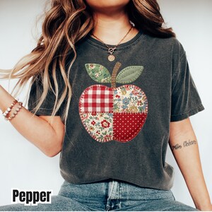 May include: Dark gray t-shirt featuring a patchwork apple design. The apple is composed of red, white, and green patterned fabrics, including gingham, floral, and polka dot patterns. The word "Pepper" is printed at the bottom of the image.