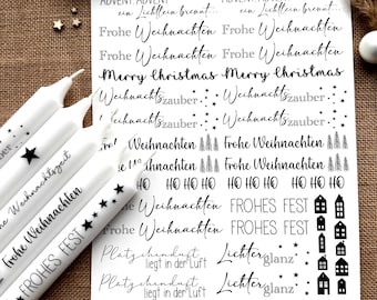 Puede incluir: Velas blancas con detalles de estrellas negras y texto que dice "Frohe Weihnachten" y "FROHES FEST". Una hoja de papel muestra frases navideñas como "Merry Christmas" y "Weihnachtszauber". Estrellas y adornos decorativos completan la escena festiva.