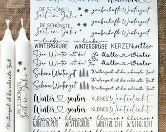 May include: Two white candles with text, next to a sheet of paper with winter-themed phrases in black script. The phrases include "Winterzeit ist die schönste Zeit" and "Zeit für Gemütlichkeit Winterlicht."