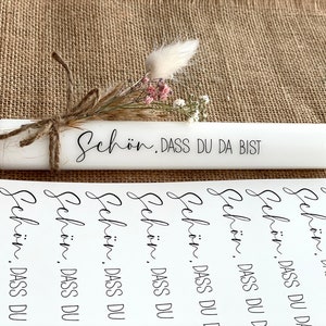 Puede incluir: Una vela blanca con el texto "Sch&ouml;n. DASS DU DA BIST" escrito en ella. La vela est&aacute; decorada con flores secas y cuerda. Hay varias hojas de papel con el mismo texto impreso en ellas.