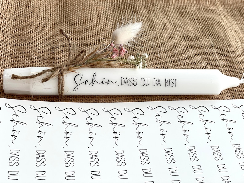 Puede incluir: Una vela blanca con el texto "Sch&ouml;n. DASS DU DA BIST" escrito en ella. La vela est&aacute; decorada con flores secas y cuerda. Hay varias hojas de papel con el mismo texto impreso en ellas.