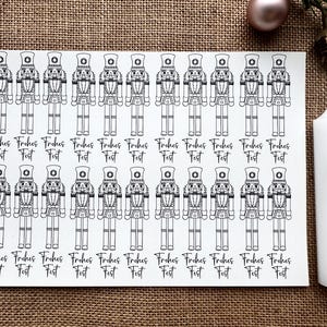 Könnte beinhalten: Ein Bogen weißer Aufkleber mit einer schwarz-weißen Illustration eines Nussknackersoldaten. Der Text "Frohes Fest" steht unter jedem Nussknacker. Eine weiße Kerze mit Nussknackermotiv ist ebenfalls zu sehen.