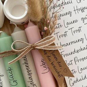 May include: Four candles with different colored wax and labels. The labels read "Gl&uuml;ck", "Gelassenheit", "Liebe", and "Timm dir, as du brauchst". The candles are tied together with twine and a small brown tag with a heart.