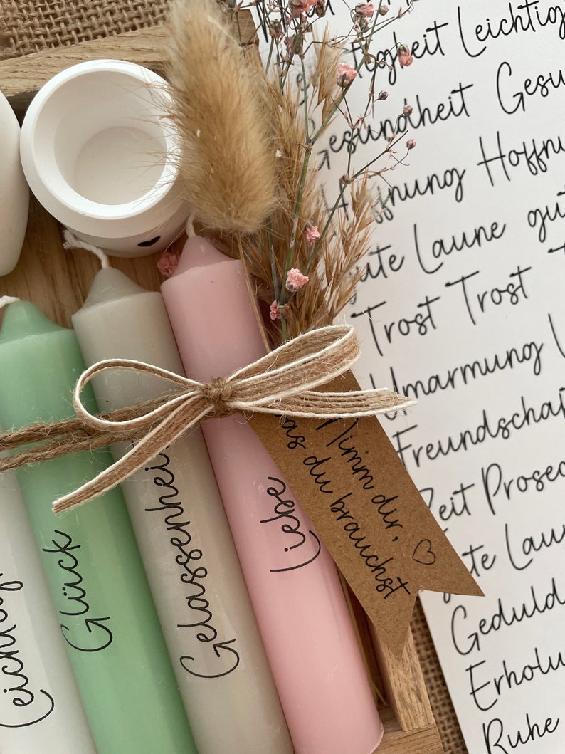 May include: Four candles with different colored wax and labels. The labels read "Gl&uuml;ck", "Gelassenheit", "Liebe", and "Timm dir, as du brauchst". The candles are tied together with twine and a small brown tag with a heart.