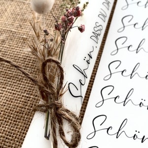 Puede incluir: Vela blanca con flores secas atadas con cuerda. La vela tiene el texto "Sch&ouml;n, dass du da bist" escrito en ella.