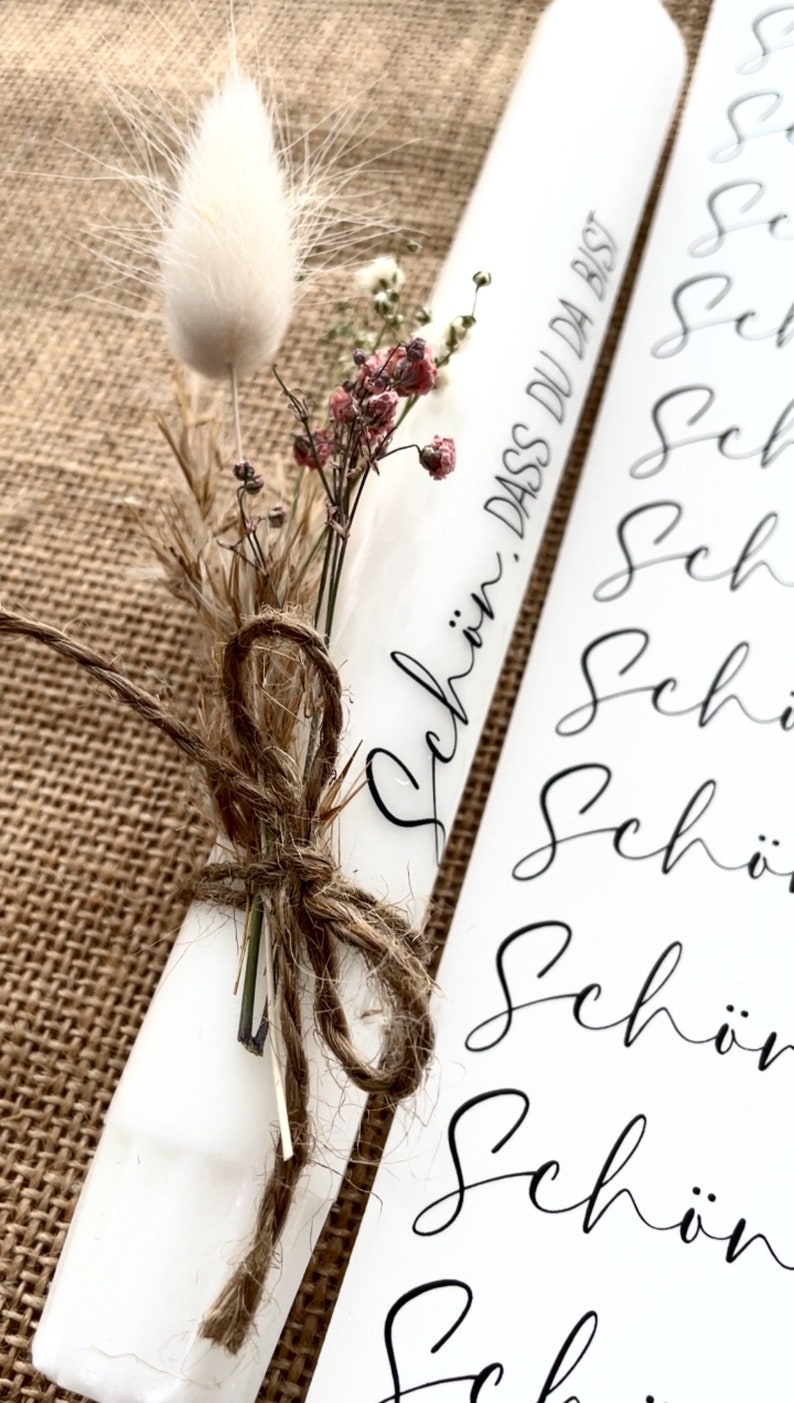 Puede incluir: Vela blanca con flores secas atadas con cuerda. La vela tiene el texto "Sch&ouml;n, dass du da bist" escrito en ella.