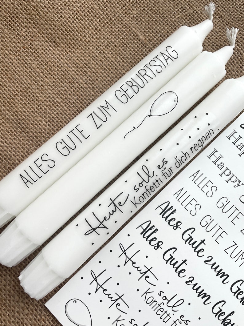 May include: Three white candles with black lettering. The candles have the text "Alles Gute zum Geburtstag" and "Heute soll es Konfetti f&uuml;r dich regnen."  The candles are decorated with black dots and a small balloon.