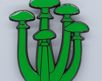 Puede incluir: Un pin de esmalte verde con un grupo de cinco setas con contornos negros. Las setas tienen sombreros redondeados y tallos.