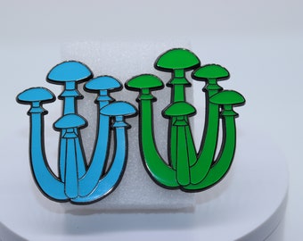 Puede incluir: Dos pines de esmalte con forma de hongo. Uno es azul claro con contorno negro, y el otro verde con contorno negro. Los pines están sobre una superficie blanca.