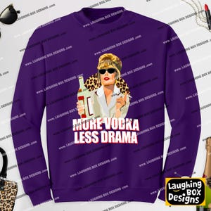May include: A deep purple long-sleeve sweatshirt featuring a graphic illustration of a person with blonde hair and sunglasses holding a bottle. Below the illustration, the text reads MORE VODKA LESS DRAMA in bold white capital letters with a red outline.