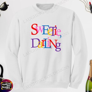 May include: A white sweatshirt with the text "Sweetie, Daring" in colorful, stacked letters. The sweatshirt is a crew neck style, and the text is in red, blue, yellow, pink, purple, and green.