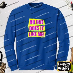 Puede incluir: Una sudadera de manga larga en color azul real con un diseño gráfico de tres rectángulos amarillos horizontales. El texto NO ONE DOES IT LIKE ME! está impreso en una fuente rosa llamativa sobre los rectángulos amarillos al frente.
