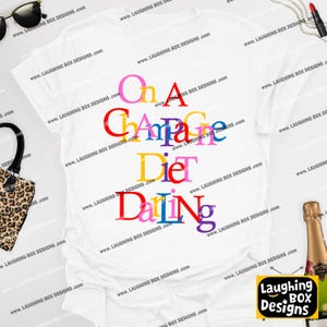 May include: White t-shirt with the text "On A Champagne Diet Darling" in colorful, stacked letters. The shirt features a V-neck and is from Laughing Box Designs.