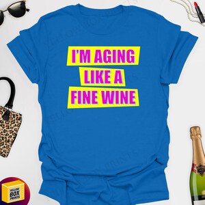 May include: A blue t-shirt with the text "I'm aging like a fine wine" in yellow and pink lettering.