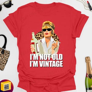 May include: A red t-shirt with a graphic of a woman in sunglasses holding a bottle and cigarette, and the text "I'M NOT OLD I'M VINTAGE". The woman is set against a leopard print background.