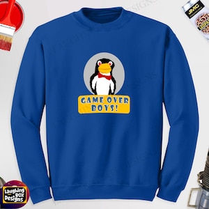May include: A royal blue sweatshirt featuring a cartoon penguin with a red bow tie, set against a gray circle. Below the penguin is a yellow banner with the text "GAME OVER BOYS!" in white. The sweatshirt is a solid color.