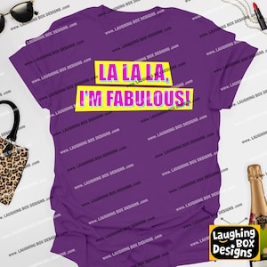 May include: A purple t-shirt with the text "LA LA LA, I'M FABULOUS!" in a yellow and blue box. The shirt is a solid color and has a crew neck. The brand name "Laughing Box Designs" is visible on the bottom right.