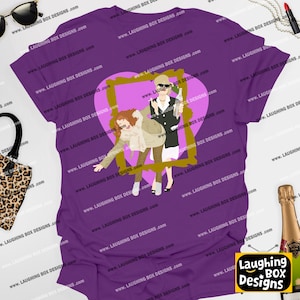 Puede incluir: Camiseta morada con una ilustración de dibujos animados de dos mujeres dentro de un marco en forma de corazón. Una mujer lleva una blazer negra y sostiene una botella, mientras que la otra lleva un abrigo color canela. El texto "Laughing Box Designs" es visible en la parte inferior derecha.