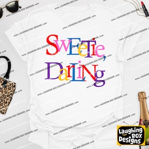 May include: White t-shirt with the words "Sweetie, Darling" in colorful block letters. The shirt features a crew neck and short sleeves. The text is in red, blue, yellow, pink, purple, and green.