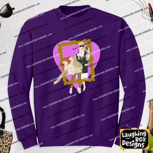 May include: A deep purple long-sleeve sweatshirt featuring a graphic print of two figures inside a gold-colored frame set against a light pink heart. The design depicts one figure in a tan coat and another in a black blazer holding a bottle.