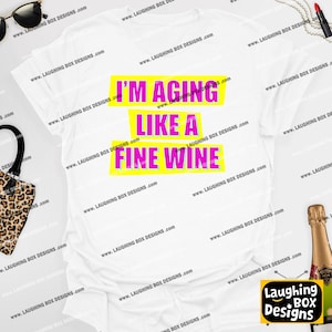May include: White t-shirt with the text "I'M AGING LIKE A FINE WINE" in pink letters on yellow rectangles. The shirt is a casual style, suitable for everyday wear. The design is centered on the front of the shirt.