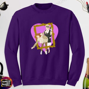 May include: A purple sweatshirt featuring a graphic of two cartoon figures inside a gold frame and a pink heart. The sweatshirt has the text "Laughing Box Designs" at the bottom.