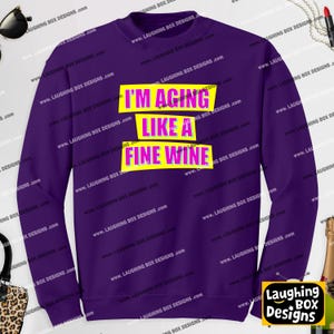 May include: A purple long-sleeve sweatshirt featuring three horizontal yellow rectangular blocks with the text "I'M AGING LIKE A FINE WINE" printed in pink capital letters across the center of the garment chest area.