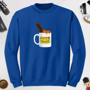 May include: Royal blue sweatshirt featuring a white mug with the text "Radio Norwich" and a graphic of beans and a sausage. The mug is filled with baked beans and a sausage is placed on top.