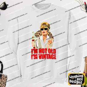 May include: A white long-sleeve sweatshirt featuring a graphic illustration of a blonde person wearing sunglasses and a leopard print scarf, holding a bottle. Below the illustration, the red text reads: I'M NOT OLD I'M VINTAGE.