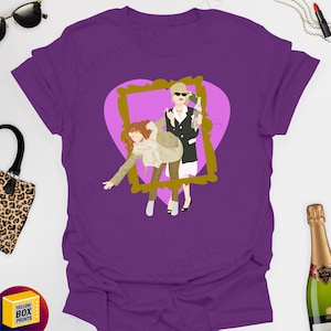 May include: Purple t-shirt featuring a pink heart with a gold picture frame. Inside the frame are two women, one holding a bottle of champagne and the other holding a glass of champagne.