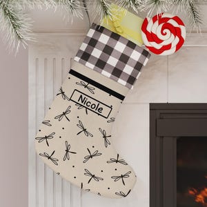 May include: A beige Christmas stocking featuring a black dragonfly pattern and a black and white buffalo check cuff. The stocking has a rectangular label with the name Nicole printed in black text, and a red and white swirl lollipop tucked inside.