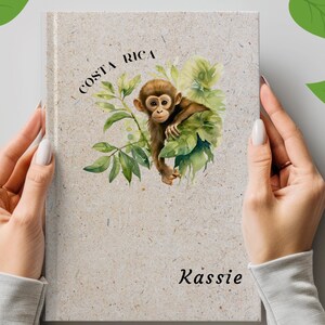 May include: A light beige, speckled notebook featuring a watercolor illustration of a brown monkey nestled among green leaves. The text "COSTA RICA" is printed in black above the monkey, and the name "Kassie" is printed in black at the bottom.