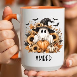 May include: A white ceramic mug with an orange handle and interior rim. The front features a graphic of a ghost wearing a black witch hat, surrounded by sunflowers, autumn leaves, and pumpkins. The name AMBER is printed in black below the design.
