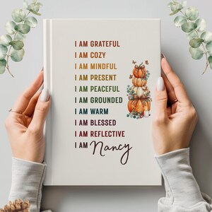 May include: A white hardcover journal featuring a stack of three watercolor pumpkins with bows and butterflies. The text reads: I AM GRATEFUL, I AM COZY, I AM MINDFUL, I AM PRESENT, I AM PEACEFUL, I AM GROUNDED, I AM WARM, I AM BLESSED, I AM REFLECTIVE, I AM Nancy.