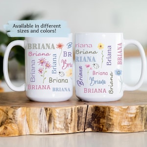 May include: Two white ceramic mugs with a floral design and the name "Briana" repeated in various colors and fonts. The mugs are on a wooden surface.