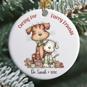 May include: A round white ceramic ornament featuring a cartoon illustration of two dogs with red and green accents, gingerbread cookies, and the text "Caring for Furry Friends" at the top and "Dr. Sarah • 2026" at the bottom with a gold string.