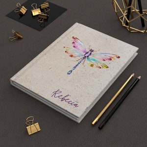 May include: A personalized journal with a watercolor dragonfly design on a beige cover. The name "Rebecca" is printed in purple on the front of the journal.