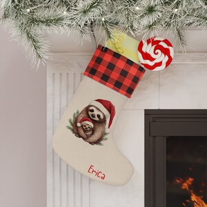 May include: A beige Christmas stocking with a red and black buffalo plaid cuff. The stocking features a watercolor illustration of two sloths, one holding the other. The stocking is personalized with the name "Erica".