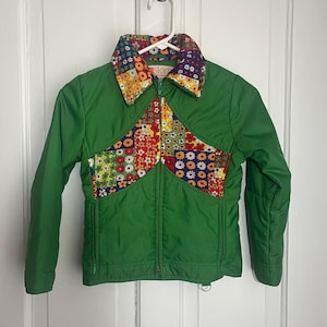 May include: A green jacket with a colorful floral patchwork design on the chest and collar. The jacket has a zipper closure and is hanging on a white hanger. The jacket is made of a shiny material.