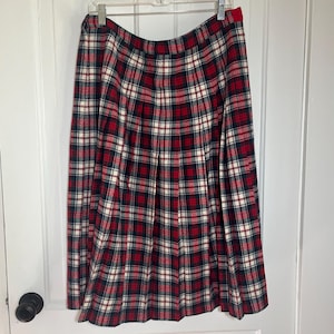 May include: A pleated plaid skirt in red, navy, and white hangs on a hanger. The skirt features a classic tartan pattern, with vertical and horizontal lines creating a grid-like design. The skirt is made of a woven fabric.