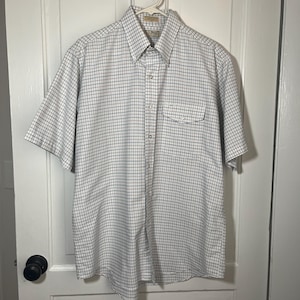 May include: A short-sleeved button-down shirt with a white and blue grid pattern. The shirt has a chest pocket and is hanging on a white hanger. The shirt is made of a lightweight fabric.