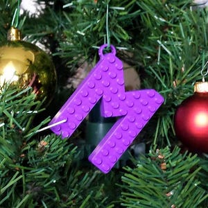 Puede incluir: Adorno navideño de color morado estilo Lego con forma de la letra M colgado de un árbol de Navidad verde.