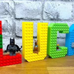 Puede incluir: Una colorida exhibición de nombre que deletrea "LUCA" hecha de grandes bloques de construcción. Las letras son rojas, amarillas, verdes y azules, con figuras de Lego de Batman, Scooby-Doo y Harry Potter. Sobre una superficie de madera.