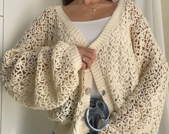 May include: A cream-colored, open-knit crochet cardigan featuring a V-neckline, long balloon sleeves, and a button-front closure. The garment is crafted with a repeating floral-inspired pattern and finished with ribbed cuffs and a ribbed hemline.