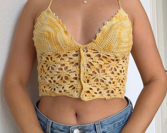May include: A yellow and white variegated crochet crop top featuring a deep V-neckline, thin spaghetti straps, and a button-down front closure. The garment is constructed with a dense stitch pattern at the bust and an open floral motif below.