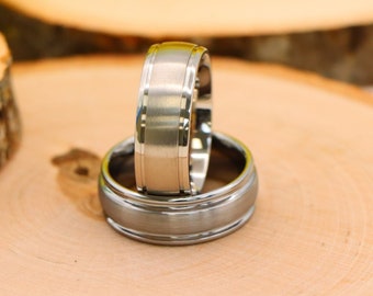 May include: Two silver wedding bands stacked on top of each other on a wooden surface.